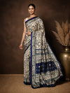 Beige printed Kashmiri silk saree with an all-over Warli-style figurative print of navy tribal figures, huts and trees, a wide navy pallu panel and a geometric navy border.